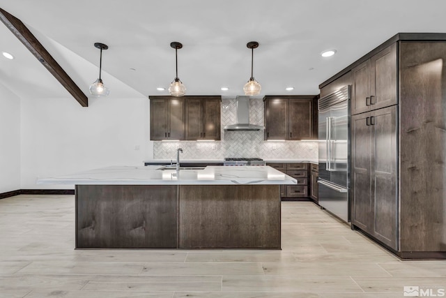 kitchen featuring wall chimney range hood, built in fridge, a sink, decorative backsplash, and dark brown cabinetry