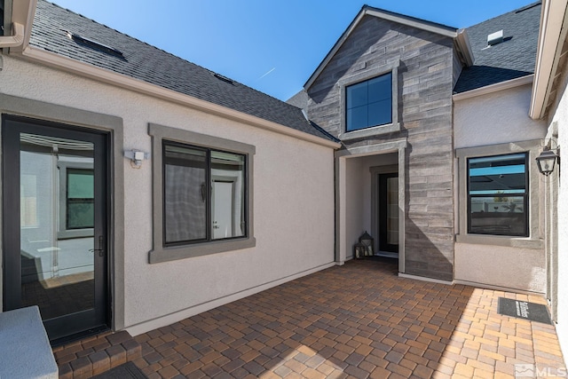rear view of house with a patio, stucco siding, roof with shingles, and stone siding