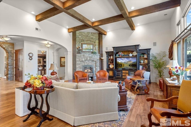 living room featuring arched walkways, beamed ceiling, wood finished floors, a stone fireplace, and a towering ceiling