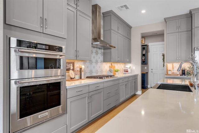 kitchen featuring stainless steel appliances, wall chimney exhaust hood, gray cabinets, a sink, and light countertops