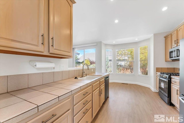 kitchen featuring appliances with stainless steel finishes, tile countertops, a sink, light wood finished floors, and light brown cabinetry
