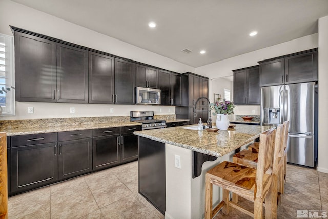 kitchen with stainless steel appliances, a sink, light stone countertops, a kitchen breakfast bar, and a center island with sink