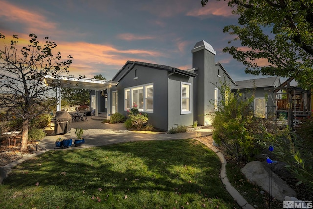 property exterior at dusk with a patio area, a lawn, stucco siding, and a chimney
