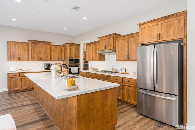 kitchen with appliances with stainless steel finishes, a sink, brown cabinets, under cabinet range hood, and dark wood-style floors