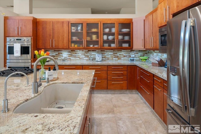 kitchen featuring stainless steel appliances, a sink, backsplash, glass insert cabinets, and brown cabinetry