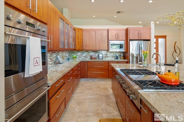 kitchen with appliances with stainless steel finishes, backsplash, light stone counters, glass insert cabinets, and brown cabinets