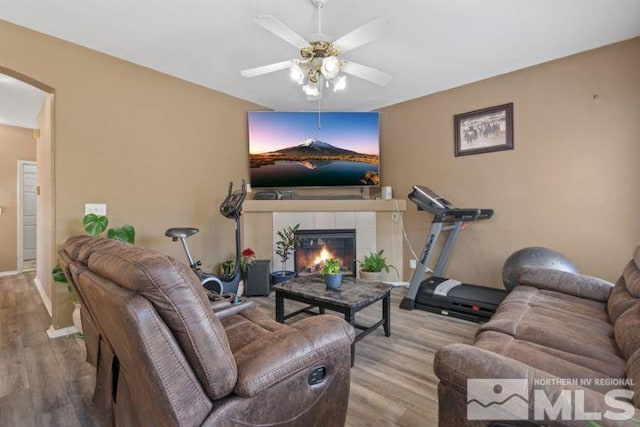 living room featuring wood finished floors, arched walkways, ceiling fan, a tiled fireplace, and baseboards
