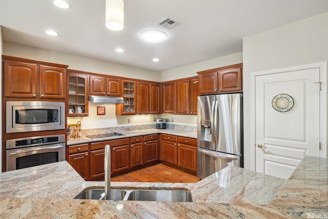kitchen with appliances with stainless steel finishes, a sink, light stone countertops, glass insert cabinets, and under cabinet range hood