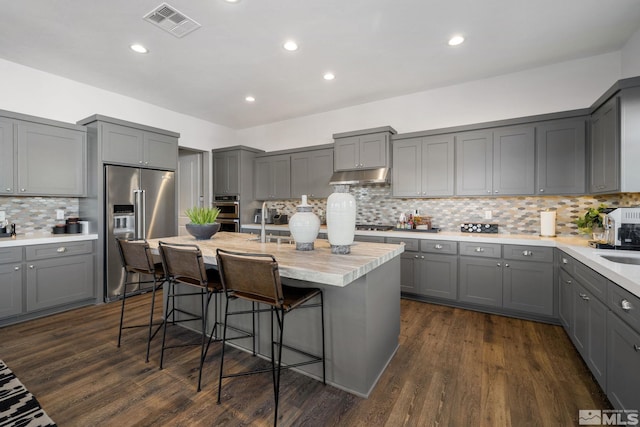 kitchen featuring gray cabinetry, high end refrigerator, under cabinet range hood, backsplash, and a kitchen breakfast bar