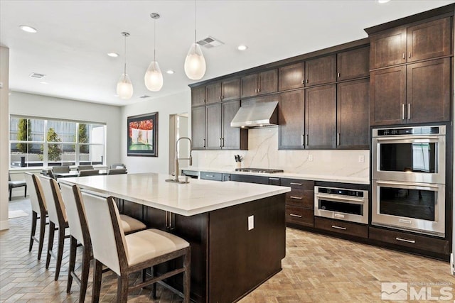 6103 S Pleasant Oak Trl, Reno, NV 89511 - kitchen featuring stainless steel appliances, brick floors, wall chimney exhaust hood, backsplash, and an island with sink