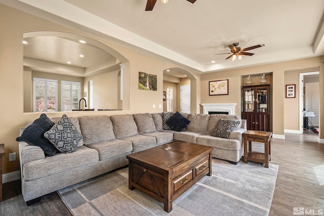 living area with a raised ceiling, a ceiling fan, arched walkways, wood finished floors, and baseboards