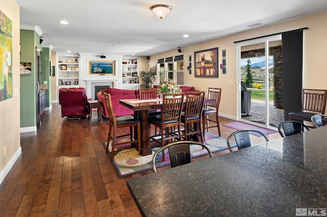 dining area featuring a warm lit fireplace, dark wood finished floors, baseboards, built in features, and recessed lighting