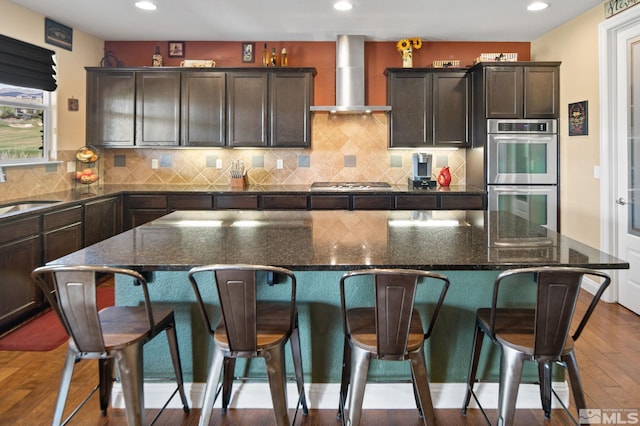 kitchen featuring wall chimney exhaust hood, appliances with stainless steel finishes, dark wood-style floors, recessed lighting, and dark brown cabinets