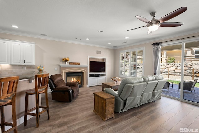 living room with wood finished floors, a lit fireplace, ornamental molding, ceiling fan, and recessed lighting
