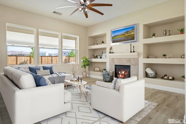 living room with built in shelves, ceiling fan, wood finished floors, a tile fireplace, and baseboards
