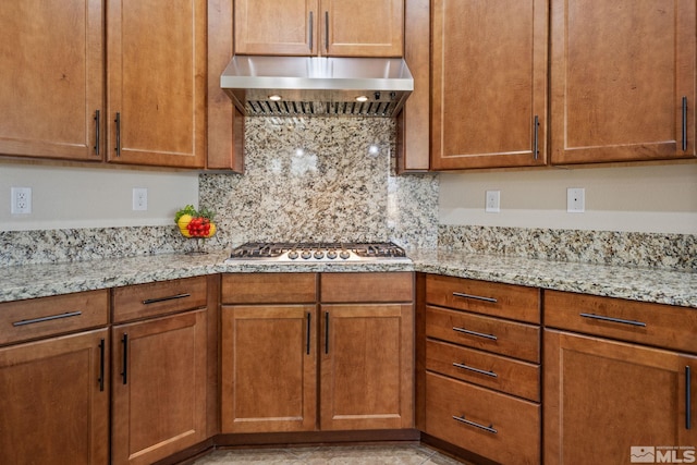 kitchen with ventilation hood, stainless steel gas stovetop, decorative backsplash, light stone counters, and brown cabinetry