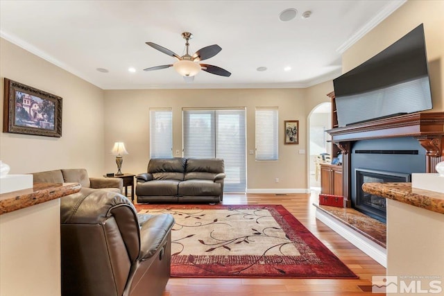 living area with ceiling fan, light wood-style floors, a glass covered fireplace, arched walkways, and crown molding