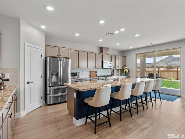kitchen with appliances with stainless steel finishes, light wood-type flooring, decorative backsplash, a center island with sink, and light stone counters