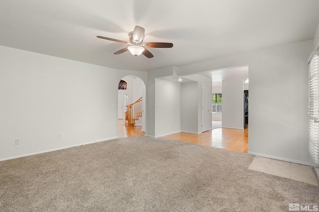 empty room with light carpet, arched walkways, ceiling fan, baseboards, and stairway