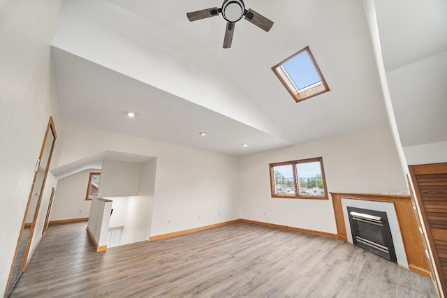 unfurnished living room featuring a skylight, light wood finished floors, a glass covered fireplace, a ceiling fan, and baseboards
