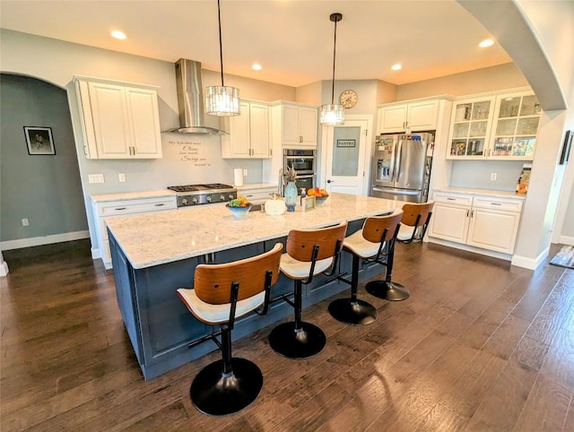 kitchen with pendant lighting, a kitchen island with sink, wall chimney exhaust hood, appliances with stainless steel finishes, and white cabinetry