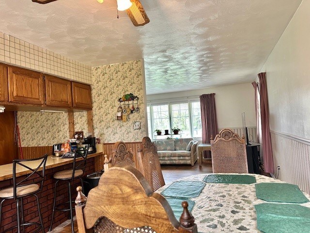 dining area with a ceiling fan, a textured ceiling, wood finished floors, a wainscoted wall, and wallpapered walls