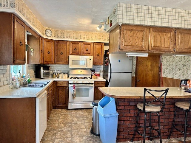 kitchen with brown cabinets, a sink, white appliances, light countertops, and a peninsula