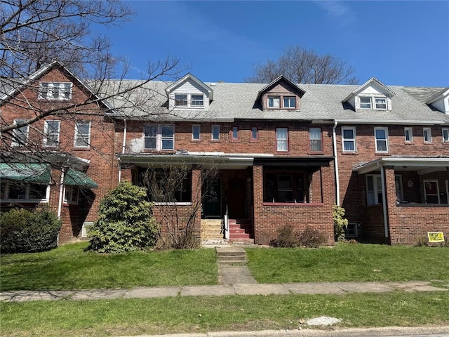 multi unit property featuring brick siding, roof with shingles, a front yard, and covered porch