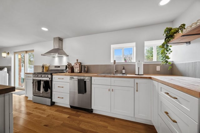 kitchen with sink, wall chimney range hood, wooden counters, white cabinets, and appliances with stainless steel finishes