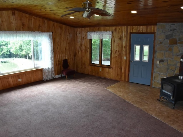 unfurnished living room featuring ceiling fan, wooden ceiling, carpet floors, a wood stove, and wood walls