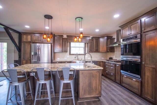 5499 Pinetree Rd, Mckean, PA 16426 - kitchen with appliances with stainless steel finishes, wall chimney range hood, a center island with sink, dark wood-style floors, and light stone countertops