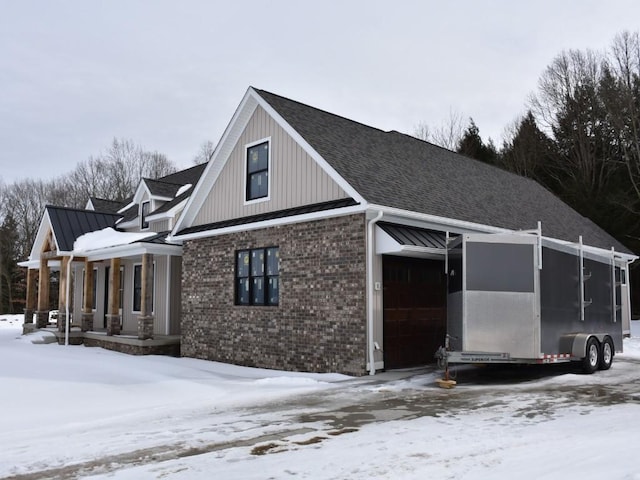 view of snow covered exterior with metal roof, a garage, brick siding, roof with shingles, and a standing seam roof