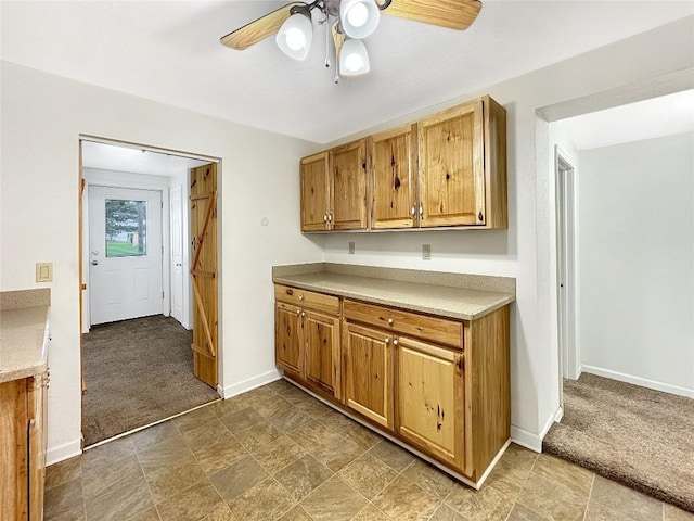kitchen featuring light countertops, stone finish floor, a ceiling fan, baseboards, and carpet flooring