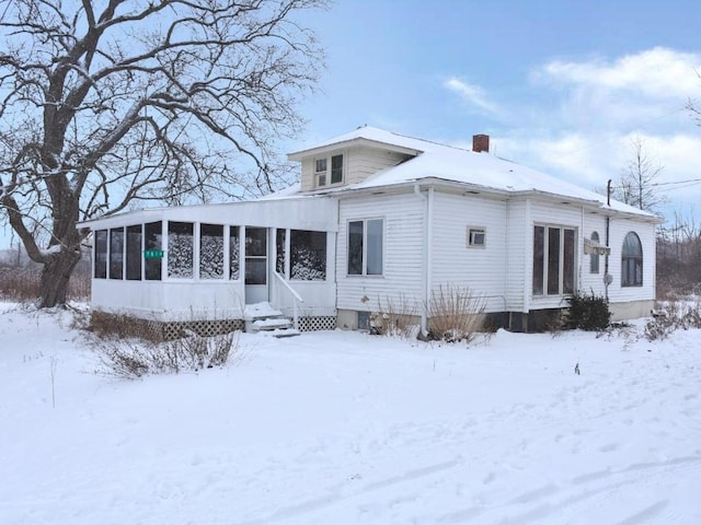 snow covered rear of property featuring entry steps, a sunroom, and a chimney