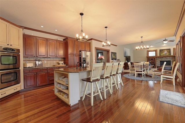 kitchen with a kitchen breakfast bar, ceiling fan with notable chandelier, stainless steel appliances, a stone fireplace, and an island with sink