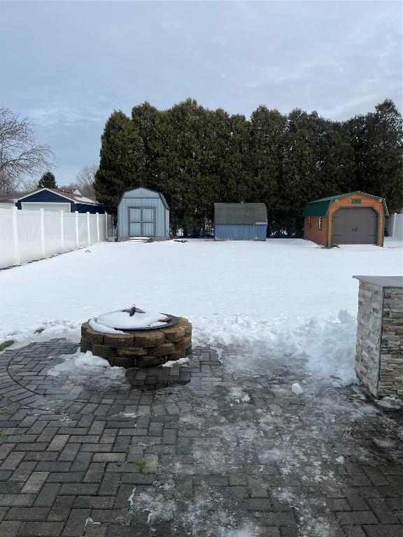 yard layered in snow featuring a garage, a fire pit, an outbuilding, fence, and a storage unit