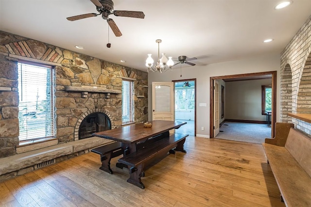 dining space with a stone fireplace, ceiling fan with notable chandelier, and light wood-type flooring