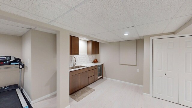 bar featuring a sink, a paneled ceiling, backsplash, baseboards, and light wood finished floors
