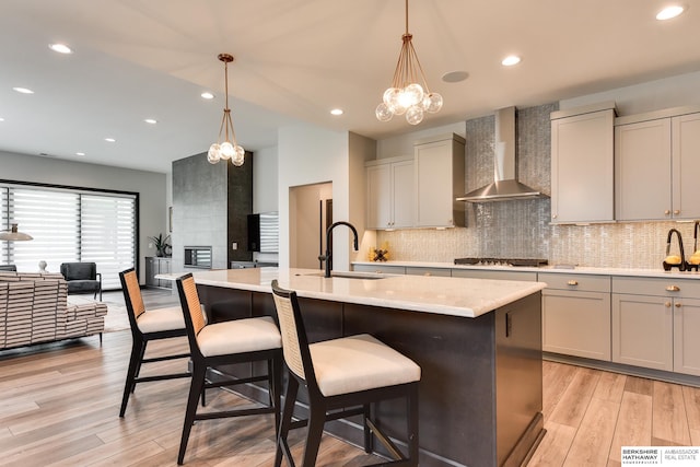 kitchen with a notable chandelier, a center island with sink, sink, light hardwood / wood-style flooring, and wall chimney exhaust hood