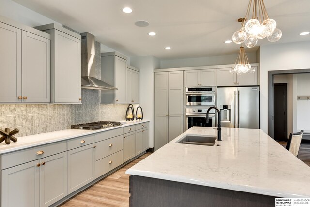 kitchen featuring a kitchen island with sink, appliances with stainless steel finishes, backsplash, wall chimney range hood, and decorative light fixtures