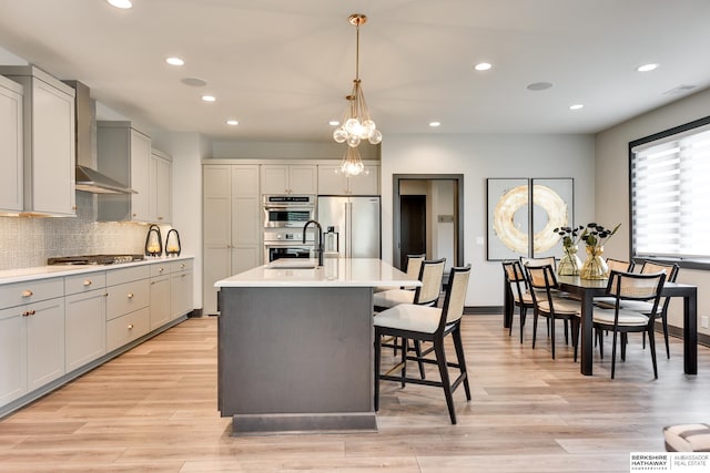 kitchen featuring decorative light fixtures, appliances with stainless steel finishes, light hardwood / wood-style floors, a kitchen island with sink, and wall chimney exhaust hood