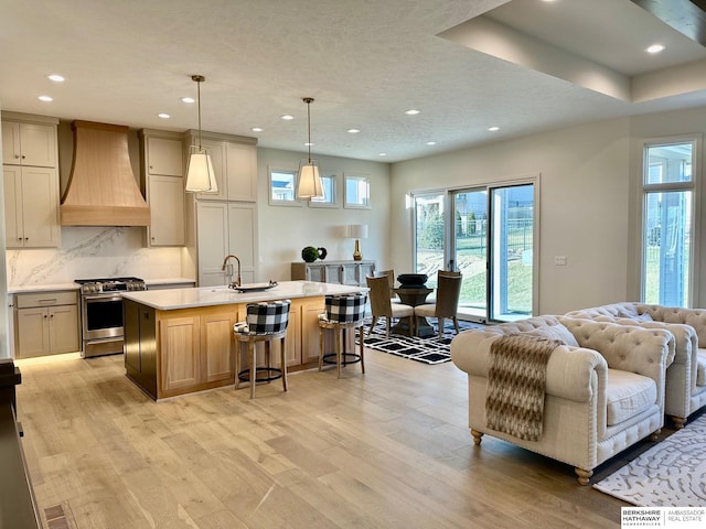 kitchen with hanging light fixtures, light hardwood / wood-style floors, custom range hood, an island with sink, and stainless steel range with gas cooktop