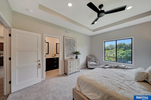 bedroom featuring light carpet, ceiling fan, ensuite bath, and a tray ceiling