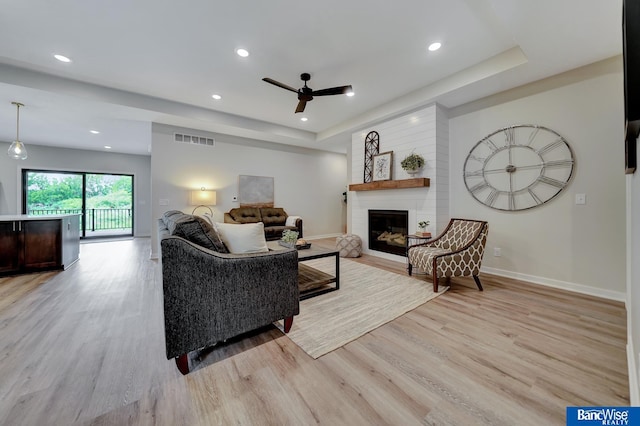 living room with ceiling fan, light wood-type flooring, a fireplace, and a tray ceiling