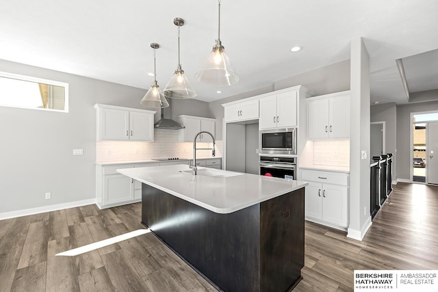 kitchen with stainless steel appliances, white cabinetry, wall chimney range hood, and a kitchen island with sink