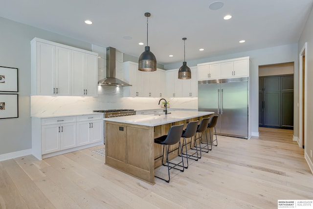 kitchen featuring wall chimney exhaust hood, white cabinetry, stainless steel built in refrigerator, a kitchen island with sink, and light wood-type flooring