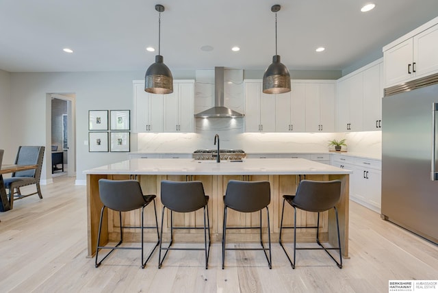 kitchen with decorative backsplash, built in refrigerator, wall chimney range hood, white cabinets, and an island with sink