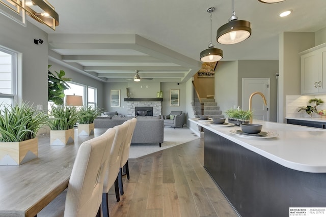 dining room with beamed ceiling, ceiling fan, a stone fireplace, and light wood-type flooring