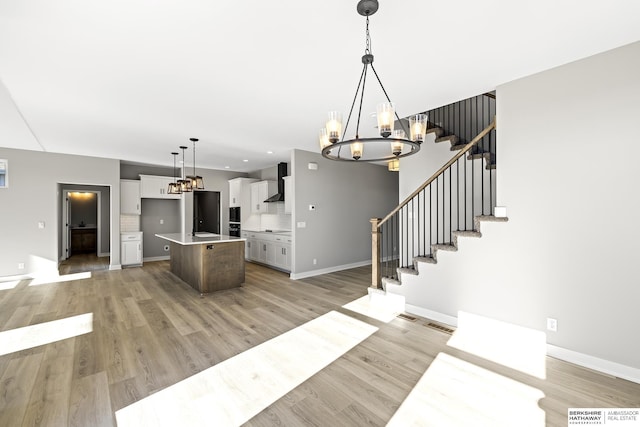 kitchen with wall chimney range hood, decorative light fixtures, decorative backsplash, a center island with sink, and light wood-type flooring