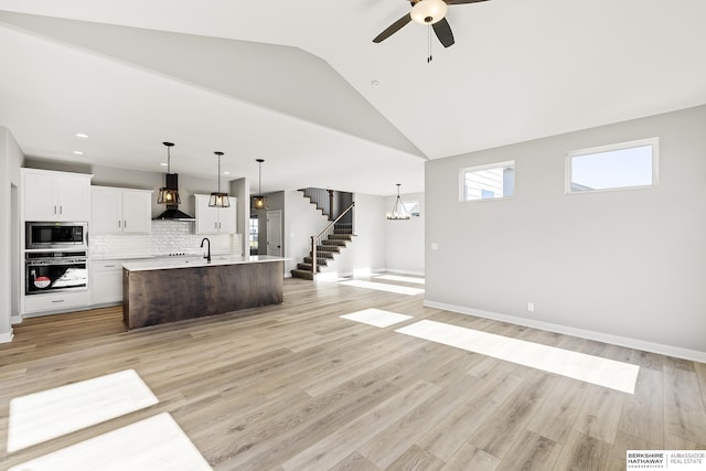 kitchen featuring stainless steel microwave, a center island with sink, wall chimney range hood, wall oven, and white cabinetry